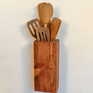 May include: A wooden utensil holder with a collection of wooden kitchen tools. The holder is a rectangular block of stained wood, and the utensils include a spoon, spatulas, and a fork. The tools are made of light-colored wood.