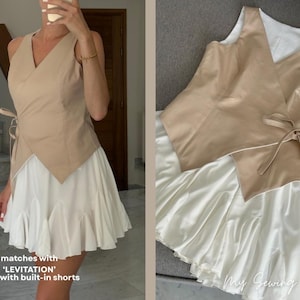 May include: A beige wrap top with a V-neck and tie closure, paired with a white skirt featuring built-in shorts. The skirt has a ruffled hem. The outfit is a modern take on a classic style.