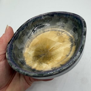 May include: A small, round ceramic bowl featuring a crystalline glaze. The center of the bowl displays a warm, golden-yellow hue with starburst-like crystal formations, while the rim transitions into a deep, dark blue and grey color palette.