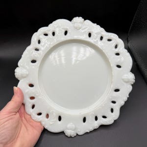 May include: A white, decorative plate with a scalloped edge and openwork design. The plate features a central, smooth surface surrounded by a detailed border with small, oval cutouts and ornate relief details. The plate is held against a dark background.