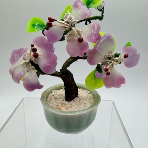 May include: A miniature bonsai tree with pink and white flower petals and green leaves. The tree is in a small, light green ceramic pot filled with white pebbles. The trunk is brown and the branches are wrapped in green wire.