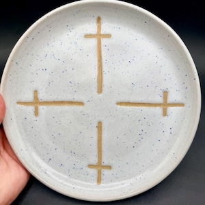 May include: A speckled white ceramic plate with four tan crosses evenly spaced around the center. The plate has a slightly raised edge and a matte finish. The plate is round and appears handmade.