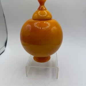 May include: A spherical orange vessel with a smooth, glossy finish and a matching lid featuring a small, rounded finial. The container rests on a circular pedestal base, showcasing a vibrant, solid color with subtle, darker orange marbling throughout.