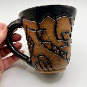 May include: A handmade ceramic mug with a black rim and handle. The body of the mug is brown with a textured, carved design in black. The mug has a unique, artistic appearance, suitable for coffee or tea.