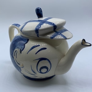 May include: A whimsical ceramic teapot with a cartoon face design. The teapot is primarily cream-colored with blue accents, including a hat-shaped lid with a plaid pattern. The spout and handle are also cream-colored.