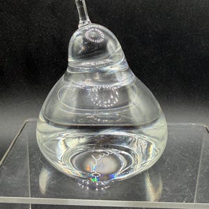 May include: A clear glass pear-shaped decorative object. The pear has a long stem and is filled with a clear liquid. The pear sits on a clear glass surface. The object is likely a decorative item for a home.