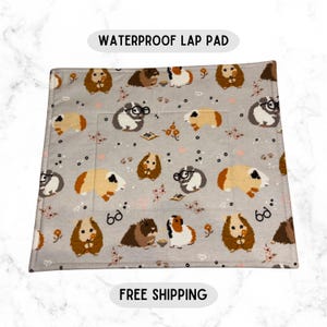 May include: A gray waterproof lap pad featuring a repeating pattern of guinea pigs in various colors and poses, some wearing glasses. The pad has a rectangular shape with a pocket and the text "WATERPROOF LAP PAD".