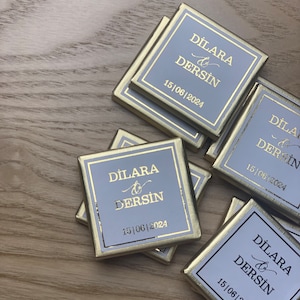 Könnte beinhalten: Ein Stapel kleiner, quadratischer, goldfarbener Schachteln mit hellblauem Hintergrund. Die Schachteln tragen den Text "DILARA & DERSIN 15|06|2024" in goldenen Lettern.