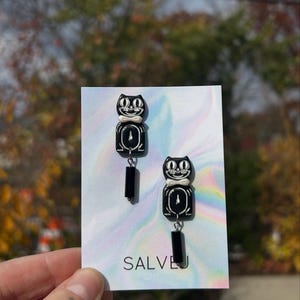 May include: A pair of black and white cat-shaped earrings. Each earring features two stacked cat faces with white bow ties and a rectangular black dangle. The earrings are displayed on a holographic card with the text "SALVEJ".