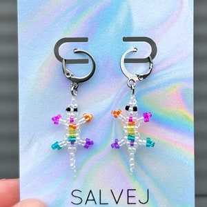 以下が含まれることがあります： シルバー色のレバーバック留めが付いた、ビーズで作られたトカゲのイヤリング。トカゲは白で、虹色のアクセントと黒い目があります。イヤリングは「SALVEJ」の文字が入ったカードに表示されています。