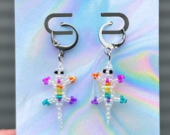 May include: Beaded lizard earrings with silver leverback clasps. The lizards are white with rainbow accents and black eyes. The earrings are displayed on a card with the word "SALVEJ".