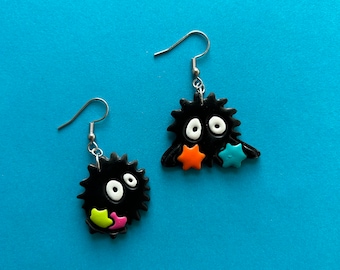 May include: A pair of black and white earrings with a fuzzy texture. Each earring features a small, round face with white eyes and a pink mouth. The earrings are decorated with colorful stars.