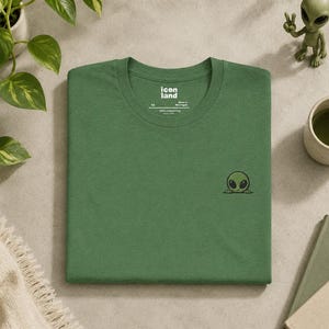 May include: A forest green t-shirt featuring a small embroidered graphic of a green alien head peeking over the bottom right corner. The inner neck label displays the text icon land, size XS, and indicates the shirt is made in Nicaragua.