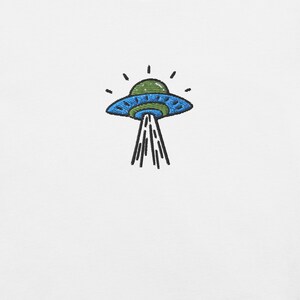 Puede incluir: Un parche bordado sobre una superficie de tela blanca que muestra un OVNI verde y azul. La nave espacial tiene una parte superior abovedada, un cuerpo ancho en forma de platillo con detalles en azul oscuro y un haz de luz blanco hacia abajo.