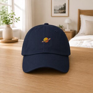 Puede incluir: Gorra de béisbol azul marino con visera curva. La gorra presenta un diseño bordado de un planeta amarillo y naranja con anillos. La gorra está hecha de una tela suave y tiene una correa ajustable.