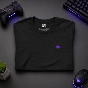 May include: A black short-sleeved t-shirt featuring a small purple pixelated alien graphic on the chest. The inner neck label displays the text icon land, size XS, and indicates the shirt is made in Nicaragua of 100% combed ring spun cotton.