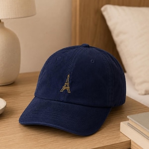 May include: A navy blue corduroy baseball cap featuring a small, centered embroidery of the Eiffel Tower in gold thread. The cap has a structured crown, a curved brim, and stitched eyelets on the top panels for ventilation and a classic fit.
