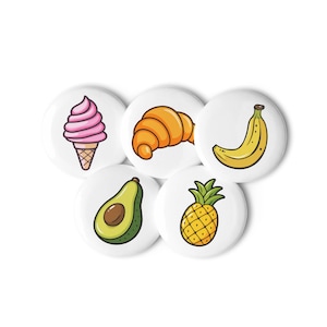 Puede incluir: Un conjunto de cinco botones redondos blancos con ilustraciones coloridas de alimentos. Los diseños incluyen un helado rosa, un cruasán dorado, un plátano amarillo, un aguacate verde y una piña amarilla. Cada botón representa un alimento diferente.