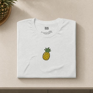 Może przedstawiać: Biały t-shirt z okrągłym dekoltem i małą, wyśrodkowaną grafiką przedstawiającą żółtego ananasa z zielonymi liśćmi. Wewnętrzna metka na karku zawiera napis icon land, rozmiar S oraz informację o produkcji w Nikaragui z 99% bawełny.
