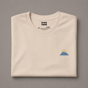 Puede incluir: Una camiseta beige de cuello redondo con un pequeño gráfico bordado en el pecho derecho de montañas azules con un sol amarillo saliendo sobre ellas. La etiqueta interior del cuello muestra el texto icon land, la talla XS y 100% combed ring.