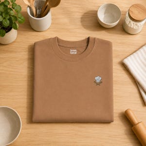 May include: A solid tan crewneck t-shirt featuring a small embroidered graphic on the chest. The embroidery depicts a white chef hat positioned above two crossed wooden spoons. The interior neck label displays the text icon land in white.
