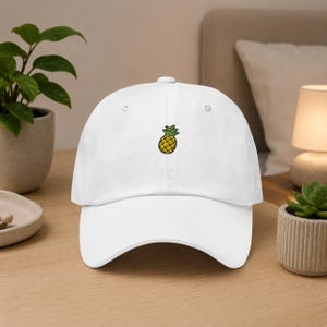 May include: White baseball cap with a small embroidered pineapple design. The pineapple is yellow with green leaves. The cap has a curved brim and a button on top. A casual accessory.