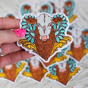 May include: Embroidered patch featuring a steer head with a white face, brown horns, and a brown beard. The steer is surrounded by turquoise and brown floral accents and yellow filigree. The patch is held in a hand.