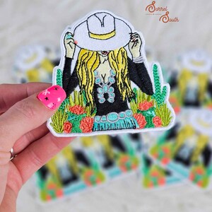 Peut inclure: Écusson brodé représentant une femme portant un chapeau de cow-boy blanc, un haut noir et des bijoux turquoise. Le motif comprend des cheveux blonds, des cactus et des fleurs rouges. L'écusson est tenu par une main avec du vernis à ongles rose.