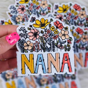 May include: Embroidered patch featuring a floral design with the word "NANA" in colorful letters. The patch showcases a variety of flowers in shades of pink, yellow, and orange, with green foliage. The word "NANA" is in yellow, blue, peach, and red.