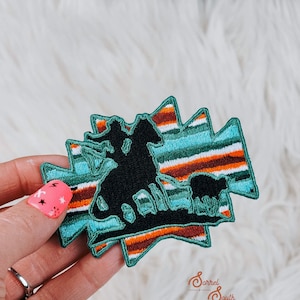 May include: A black embroidered patch with a silhouette of a cowboy riding a horse and a cow. The patch is on a turquoise and orange striped background. The text "Barrel South" is visible in the bottom right corner of the image.