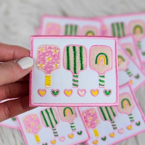 May include: Embroidered patch featuring three colorful paddle designs. The paddles are pink, green, and peach, with floral, striped, and rainbow patterns. Yellow, green, and pink hearts are below the paddles. The patch has a white background and a pink border.