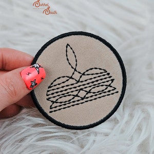 May include: A beige round embroidered patch with black thread depicting a stack of pancakes with a single flame on top. The patch has a black border.