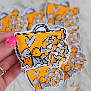 May include: A yellow embroidered patch with a heart, a bow, and a pom-pom. The patch is designed to look like a cheerleading bag.
