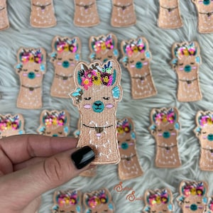 May include: Embroidered llama patches in various sizes. The llama has a tan body, a colorful floral crown, and a turquoise nose. The patches are on a white, fluffy surface. One patch is held by a hand with a black fingernail.