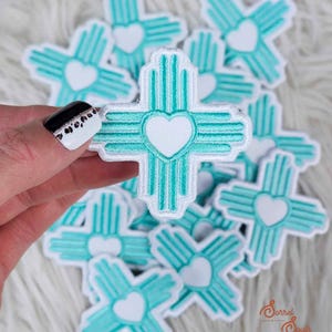 May include: A collection of turquoise and white embroidered patches in the shape of a cross with a heart in the center. The patches are on a white, fluffy surface. The patches have a Southwestern design.