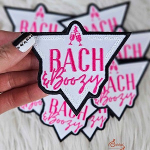 Könnte beinhalten: Ein Set mit fünf weißen, sternförmigen Bügeletiketten mit schwarzen Umrandungen. Die Etiketten zeigen den Text "Bach & Boozy" in rosa Schrift mit einer rosa Champagnerglasgrafik.