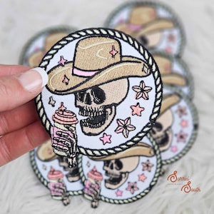 May include: A white embroidered patch with a cartoon skull wearing a brown cowboy hat with a pink band. The skull is holding a pink and white cup with a straw. The patch is surrounded by a black rope border and has pink stars and flowers.