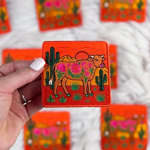 Puede incluir: Parche bordado cuadrado con un diseño de vaca adornada con motivos florales, sobre un paisaje desértico con cactus y una puesta de sol. El parche tiene un fondo naranja y costuras detalladas. Se ven otros parches con el mismo diseño.