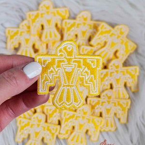 Może przedstawiać: Żółto-biała, haftowana naszywka z wystylizowanym wzorem ptaka. Naszywka ma szczegółowy kontur i jest trzymana przed stosem podobnych naszywek. Naszywki są prawdopodobnie przeznaczone na odzież lub akcesoria.