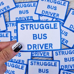 Könnte beinhalten: Eine Sammlung weißer Stoff-Patches mit blau gesticktem Text, der "Struggle Bus Driver" lautet.