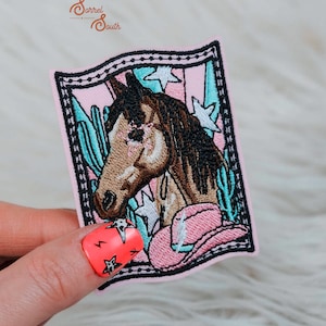 May include: Embroidered iron-on patch featuring a brown horse with a pink cowboy hat, surrounded by turquoise cacti and white stars on a pink background. The patch is framed with a black and white border.