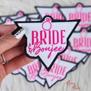 Pode incluir: Um remendo de ferro em forma de diamante, com tecido branco e guarnição preta. O remendo diz "Bride & Boujee" em letras cor-de-rosa, com um gráfico de anel de diamante cor-de-rosa acima do texto.