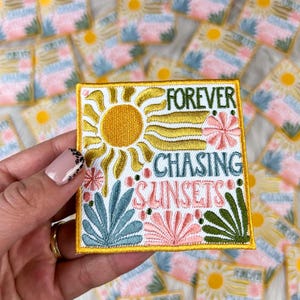 Puede incluir: Parche bordado con las palabras "FOREVER CHASING SUNSETS" en verde y azul. El parche presenta un sol amarillo, flores rosas y hojas azules. El parche es cuadrado con un borde amarillo.
