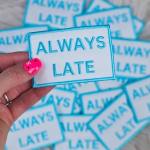 May include: A close-up of a hand holding a white patch with blue embroidered text that reads "ALWAYS LATE". The patch is surrounded by other similar patches.