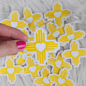 May include: A pile of yellow and white embroidered iron-on patches featuring the symbol of the state of New Mexico with a heart in the center.