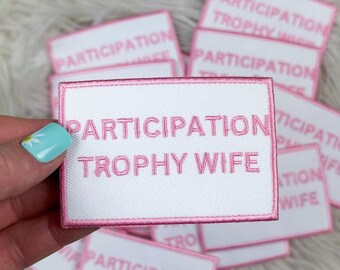Puede incluir: Un parche blanco con costuras rosas y el texto "Participation Trophy Wife" en letras rosas.