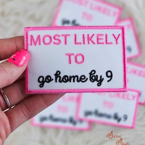 May include: A white rectangular patch with pink embroidered text that reads "MOST LIKELY TO go home by 9". The patch has a pink border and is held in a hand. Other patches are blurred in the background.