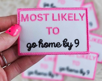 Puede incluir: Un parche rectangular blanco con texto bordado en rosa que dice "MOST LIKELY TO go home by 9". El parche tiene un borde rosa y se sostiene en una mano. Otros parches están borrosos en el fondo.