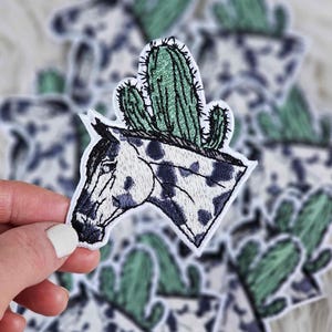 May include: Embroidered patch featuring a horse head with a cactus on top. The horse has a white base with dark blue spots. The cactus is green with black detailing. The patch is white with a black border. The patches are scattered on a white surface.