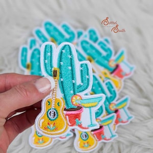 May include: A set of embroidered iron-on patches featuring a cactus with a guitar, a margarita, and a lemon slice. The patches are white with colorful embroidery.